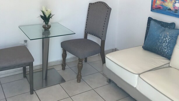 Un salón con una mesa de vidrio, un sillón tapizado en gris, un sofá blanco con un cojín azul y dos cuadros en las paredes.
