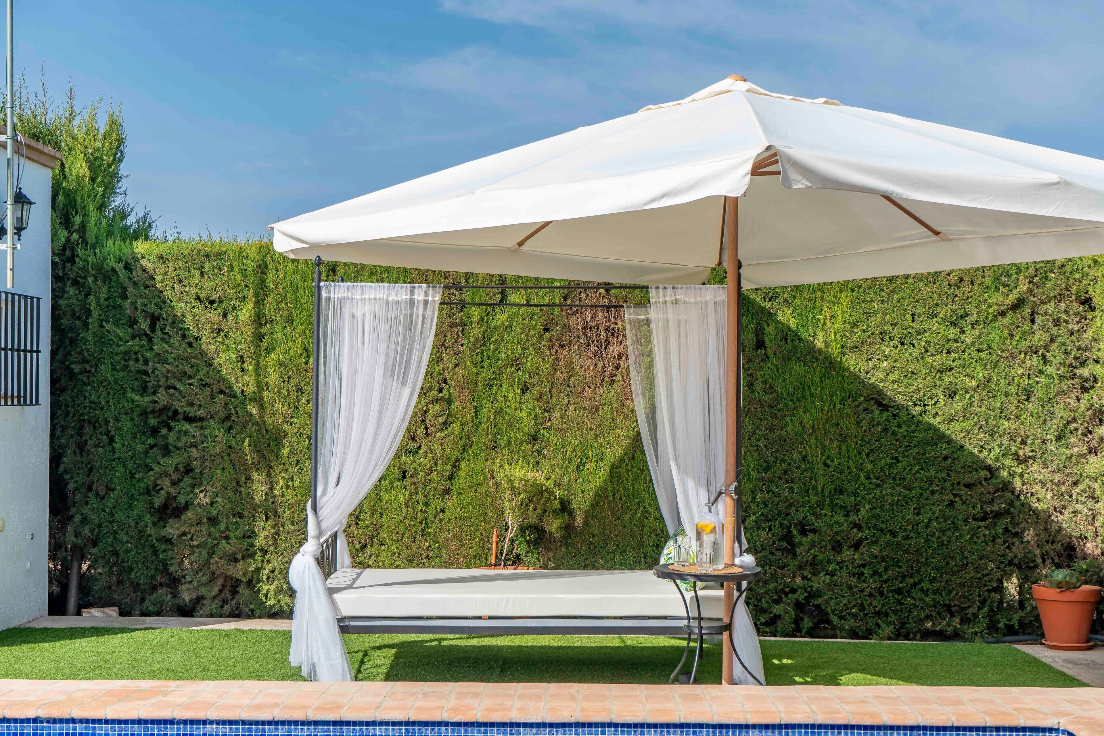 A poolside area with a white canopy, a small table with a bottle and glass, and a neatly trimmed hedge in the background.