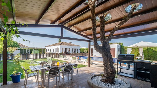 Une terrasse extérieure avec une table à manger, des chaises, un barbecue et une piscine en arrière-plan.