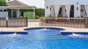 Une piscine avec une terrasse en bois, un patio couvert et un bâtiment blanc doté d’un toit en tuiles.