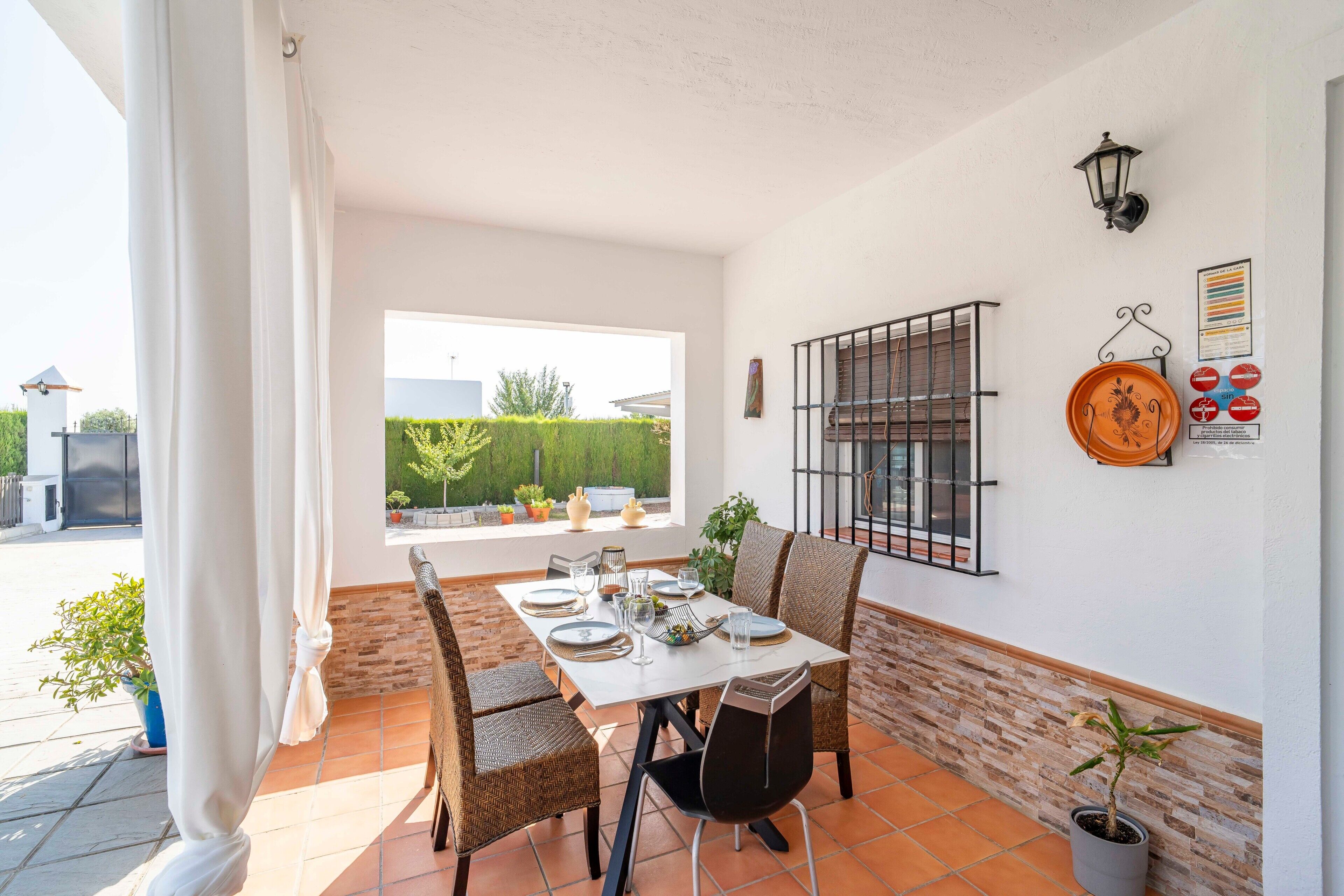 Comedor con una mesa preparada para cuatro personas, sillas de mimbre, una planta en maceta y vistas al exterior.
