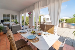 Un área de comedor al aire libre con una mesa blanca para dos personas, mobiliario de mimbre y vistas a una piscina y follaje.