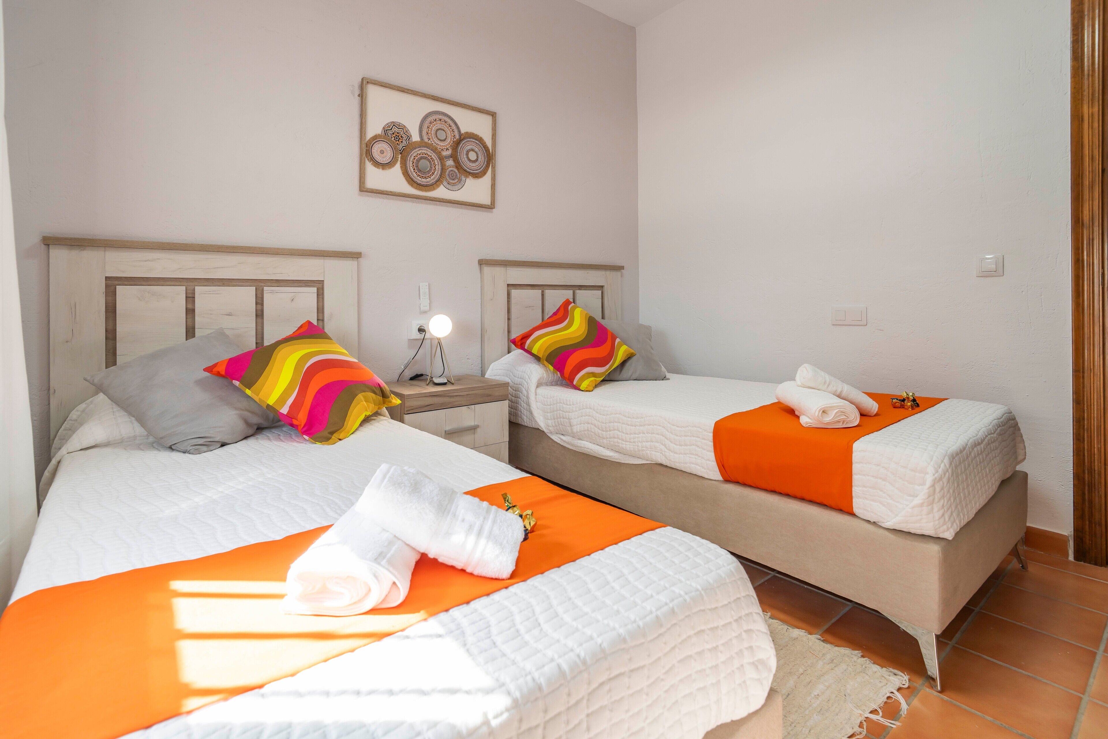 A hotel room with two beds, each with a white bedspread and orange accents, a bedside table with a lamp, and a wall-mounted artwork.