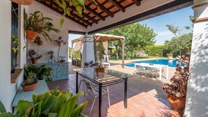 An outdoor patio area with a glass table, chairs, potted plants, a pergola, and a pool in the background. - Rural House 'Carula' with Private Pool, Wi-Fi and Air Conditioning (La Puebla de Cazalla)