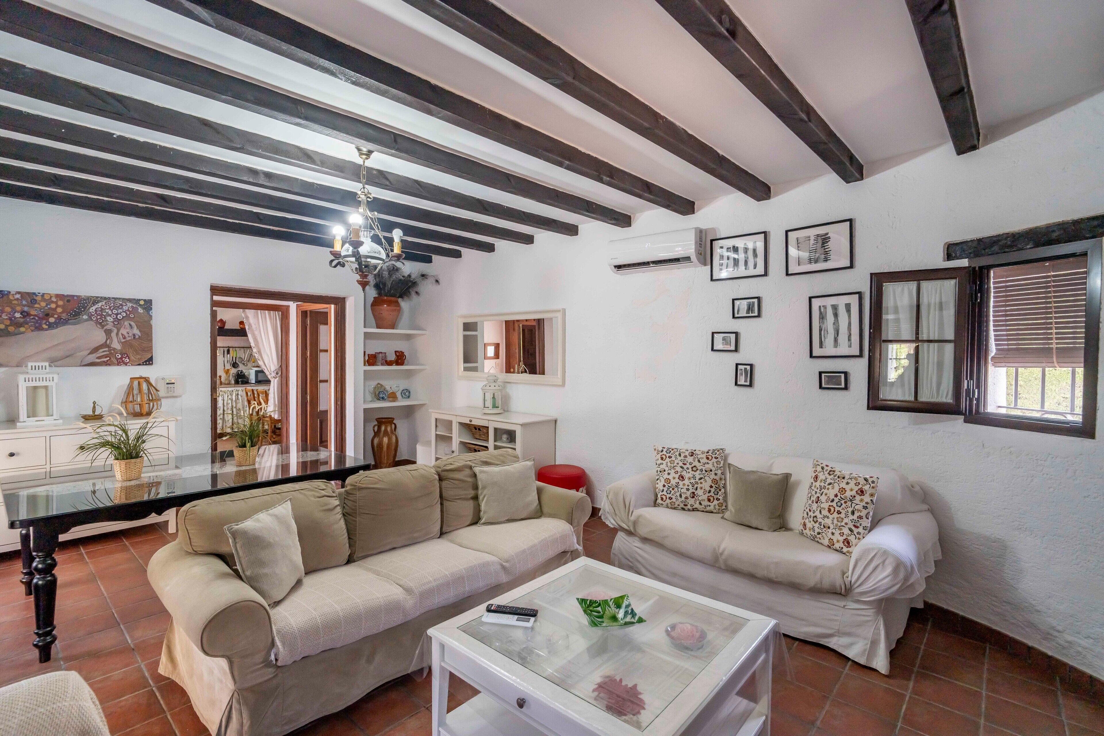 A living room with a sofa, a coffee table, and a TV. There are shelves with decorative items and a window with blinds.