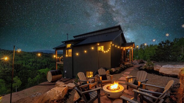 A cozy outdoor seating area with Adirondack chairs and a fire pit, set against a backdrop of a starry night sky and a small cabin with string lights. - Golden Heights - Sauna + 180 Degree Views + Hot Tub (Cripple Creek)