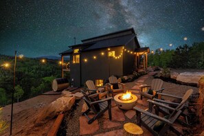 Un espace extérieur accueillant, aménagé avec des fauteuils Adirondack et un foyer, le tout mis en valeur par un ciel étoilé et une petite cabane illuminée de guirlandes lumineuses.