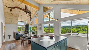 A spacious living area with a large island, wooden ceiling, and large windows offering a view of a forested landscape. - Golden Heights - Sauna + 180 Degree Views + Hot Tub (Cripple Creek)