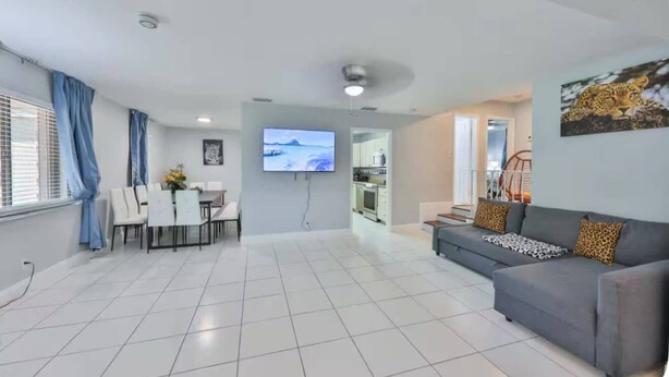 A spacious living area with a grey sofa, a dining table with chairs, a flat-screen TV mounted on the wall, and a kitchen visible through an open doorway.