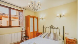 A bedroom with a bed, a wooden wardrobe, a nightstand, a chair, a chandelier, and a window with curtains.