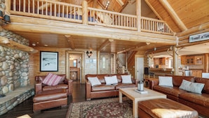 A spacious log cabin interior with a high ceiling, a stone fireplace, leather sofas, and a wooden staircase. - Northern Exposure Lakefront (Fawnskin)