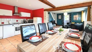 Un comedor con una mesa de madera dispuesta para cuatro personas, rodeada de sillas de cuero negro. Se ve una cocina con armarios blancos y una pared roja al fondo.