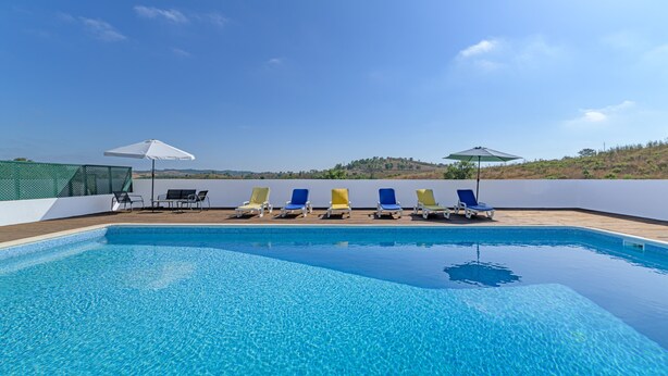 Piscina com espreguiçadeiras e guarda-sóis, cercada por um muro branco e vista para colinas.