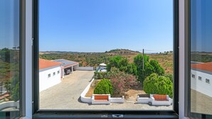 Vista pela janela de um edifício com um pátio pavimentado, rodeado por vegetação e uma paisagem montanhosa.