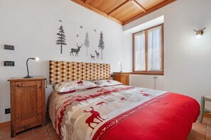 A bedroom with a checkered headboard, a bed with a red and beige patterned bedspread, a wall-mounted lamp, a window with curtains, and a wall decal of animals and trees.