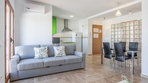 A modern living room with a grey sofa, a dining table with chairs, a kitchen in the background, and a decorative wall panel.