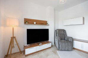 A modern living room with a flat-screen TV, a grey armchair, a wooden shelf with decorative items, a floor lamp, and a white cabinet. - Apartment 'Viento Del Mar' with Shared Pool, Private Terrace and Air Conditioning. (Chiclana de la Frontera)