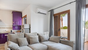 Un salon moderne avec un canapé beige, un petit coin cuisine doté de meubles violets et une porte-fenêtre coulissante donnant sur un espace extérieur.