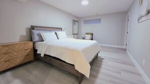 A bedroom with a bed, a nightstand, a dresser, a ceiling light, a window, and a wall-mounted mirror. - 🏡 Spacious Basement Suite: 2 queen beds, Kitchen, Livingroom, Dining, Laundry (Edmonton)