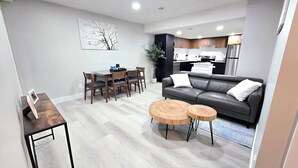 A modern living room with a sofa, two wooden side tables, a dining table with chairs, and a kitchen in the background. - 🏡 Spacious Basement Suite: 2 queen beds, Kitchen, Livingroom, Dining, Laundry (Edmonton)