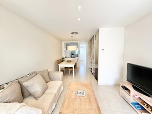 A living room with a sofa, coffee table, TV, and a dining area in the background.