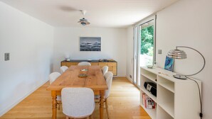 Un comedor con una mesa y sillas de madera, una cocina al fondo y vistas a través de grandes ventanales.