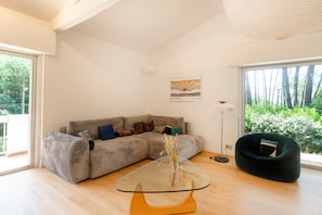 A modern living room with a sectional sofa, a glass coffee table, a dark armchair, and a large window with a view of trees. - Villa Estrella between lake and ocean with private pool, private terrace, and Wi-Fi (Soorts-Hossegor)