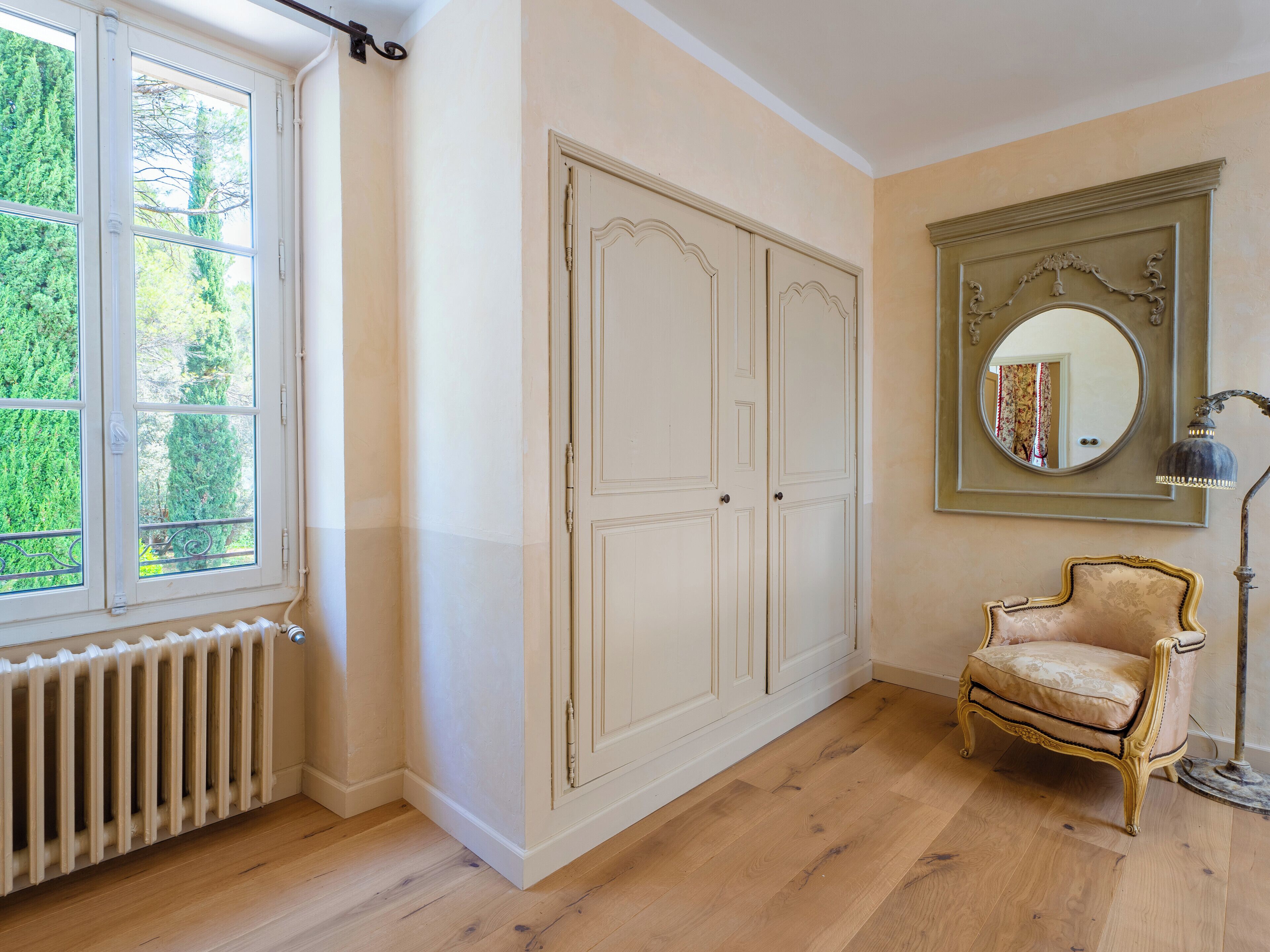 Ein Zimmer mit Heizkörper, Fenster mit Blick auf Bäume, ein weißer Kleiderschrank, ein Stuhl und ein Spiegel mit Lampe.