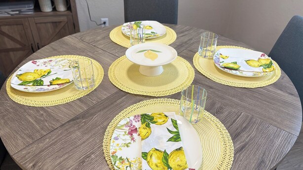 Uma mesa de jantar redonda de madeira, com jogos americanos amarelos, pratos com motivos florais e copos de vidro transparente.