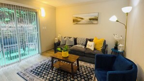 Uma sala de estar com sofá, mesa de centro, poltrona azul, tapete estampado e um quadro na parede.