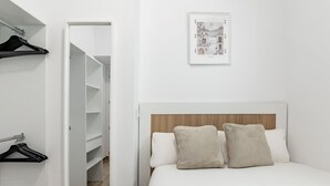 A bedroom with a bed, a wooden headboard, a white wall, a framed picture, and a closet with hanging clothes. - San Isidro IV - Cozy apartment (Madrid)