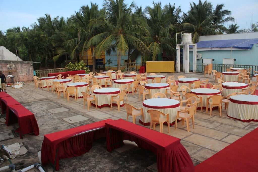 Une terrasse extérieure avec des tables et des chaises rondes, des palmiers en arrière-plan et un espace scène avec des rideaux rouges.
