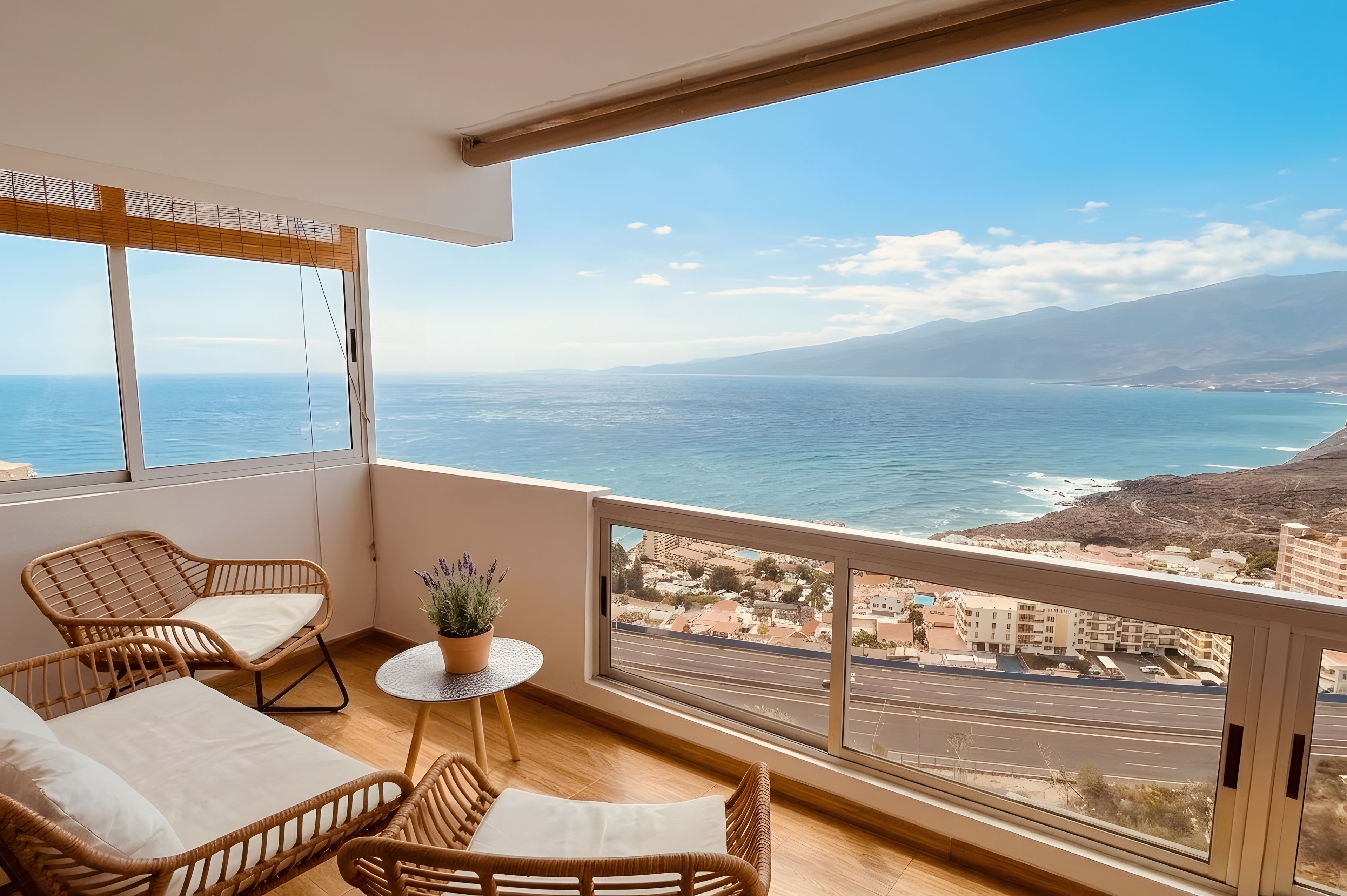 A balcony with wicker furniture, a small table, and a potted plant, overlooking a coastal view with buildings and a road.