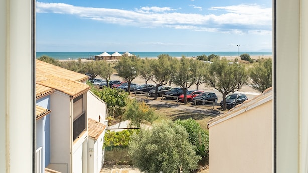 A view of a beachside parking area with several cars, a row of trees, and a building with a tiled roof. - Apartment 'Les Balcons De La Méditerranée' with Sea View and Balcony (Narbonne-Plage)