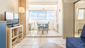 A room with a television on a stand, a dining table with chairs, a sofa, and a view of the sky through a window. - Apartment 'Les Balcons De La Méditerranée' with Sea View and Balcony (Narbonne-Plage)