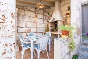 Un espace de restauration extérieur de style rustique, avec un mur en pierre, des chaises en osier blanches, une table en verre, des plantes suspendues et une cheminée.