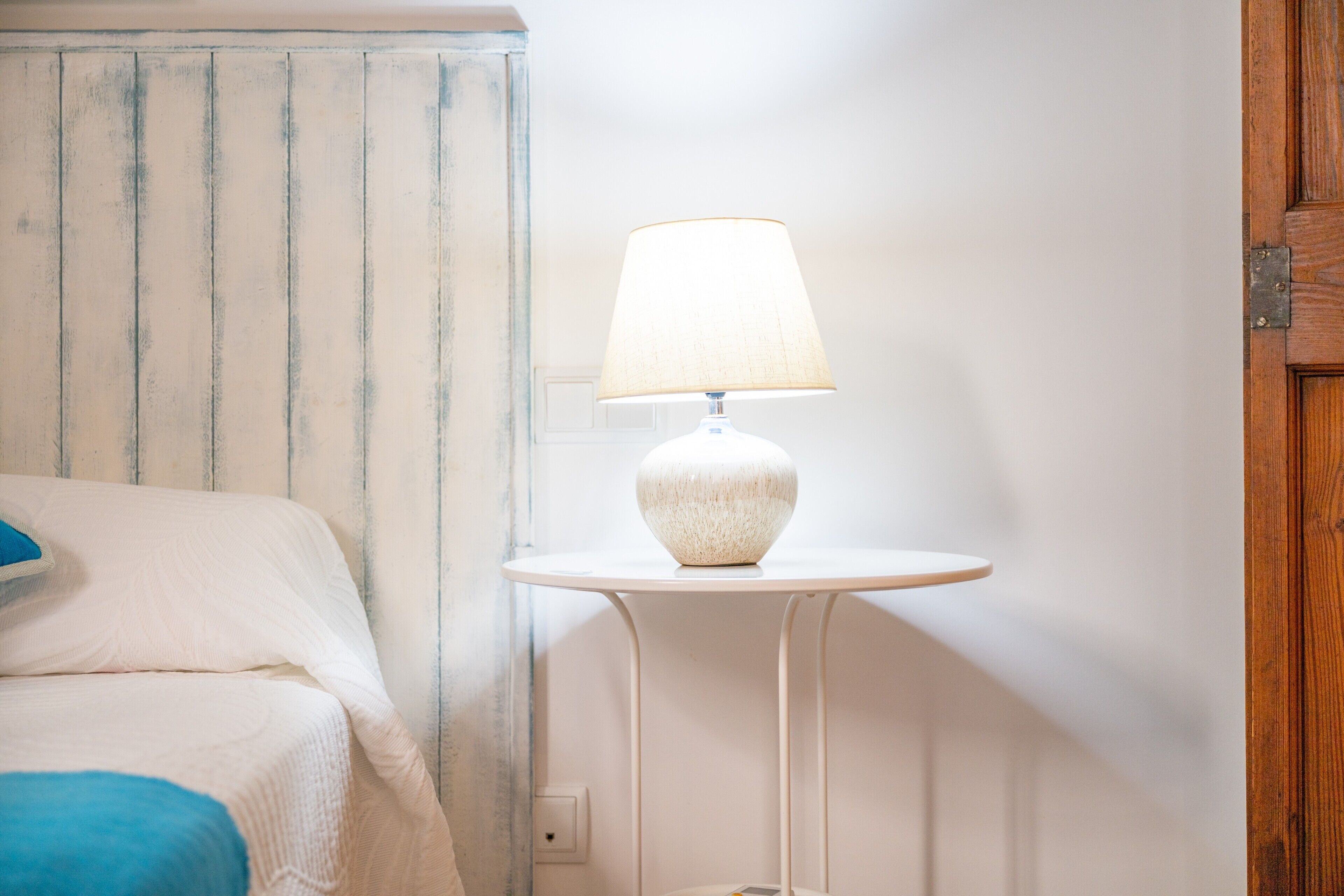 Una cama con un almohadón azul, una lámpara blanca sobre una mesita y una puerta de madera.