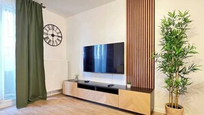 A modern living room with a flat-screen TV mounted on a wall, a wooden TV stand, a large potted plant, and a clock on the wall. - T2D Apartment - Suite Gallery Disneyland Paris (Lognes)
