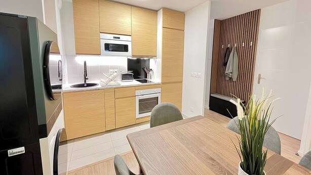A modern kitchen with wooden cabinets, a microwave, and a sink. There is a dining table with chairs and a potted plant. - T2D Apartment - Suite Gallery Disneyland Paris (Lognes)