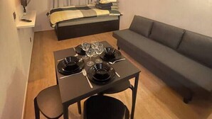 A compact room with a bed, a sofa, a dining table with chairs, and a small table with wine glasses and plates. - T2D Apartment - Suite UR near Disneyland Paris (Bussy-Saint-Georges)