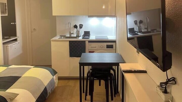 A compact hotel room with a kitchenette, a bed with a checkered duvet, a small dining table, and a TV mounted on the wall. - T2D Apartment - Suite UR near Disneyland Paris (Bussy-Saint-Georges)
