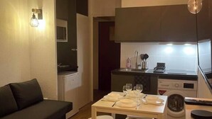 A compact living space with a kitchenette, a dining area with a table set for two, and a dark-colored sofa. - T2D Apartment - Suite BLG near Disneyland Paris (Bussy-Saint-Georges)