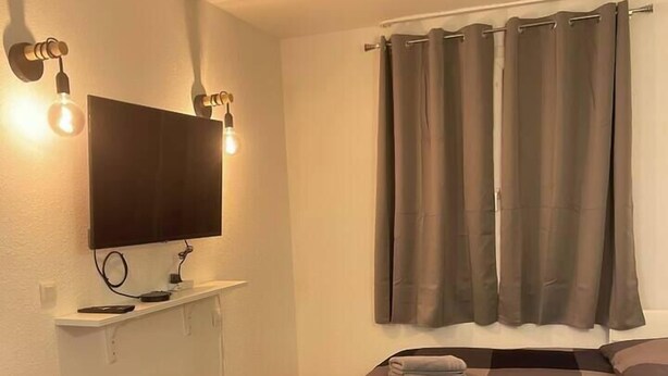 A bedroom with a bed, a TV mounted on the wall, a small table, and a window with curtains. - T2D Apartment - Suite BLG near Disneyland Paris (Bussy-Saint-Georges)