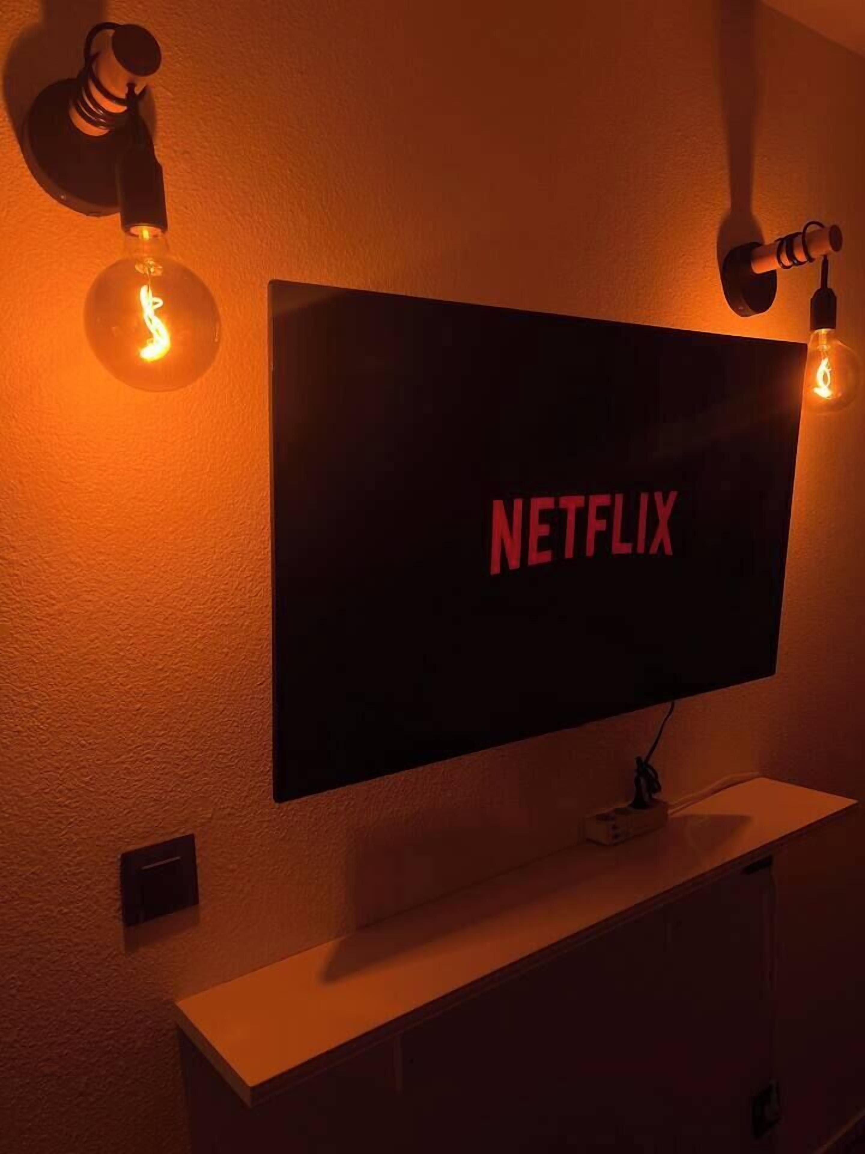 Ein an der Wand befestigter Fernseher mit dem Netflix-Logo, umgeben von zwei Wandleuchten mit Glühbirnen.