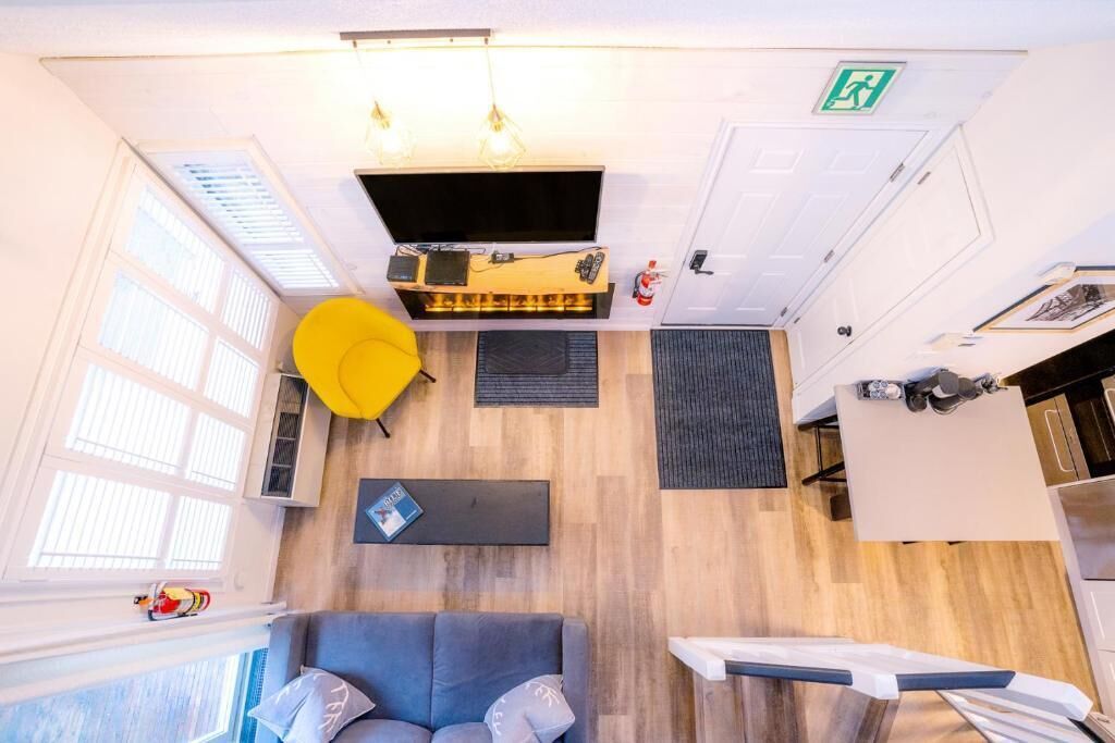 Habitación de hotel moderna con sofá, una silla amarilla, una pequeña mesa y una zona de cocina.