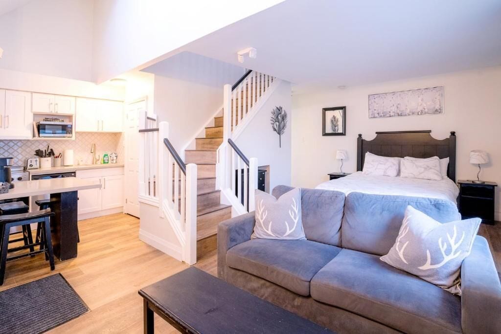 A compact living space with a kitchenette, a sofa with decorative pillows, a bed with a headboard, and a staircase.