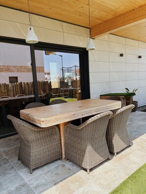An outdoor patio with a wooden table and wicker chairs, a wooden ceiling, and a view of the surrounding area.