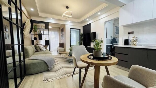 Un salon moderne comprenant un canapé, des fauteuils, une table basse et un téléviseur. Il y a également un coin cuisine avec des placards et un plan de travail.