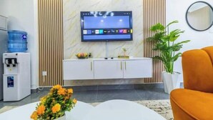 A modern living room with a flat-screen TV mounted on the wall, a white cabinet with gold handles, a water dispenser, and a potted plant.