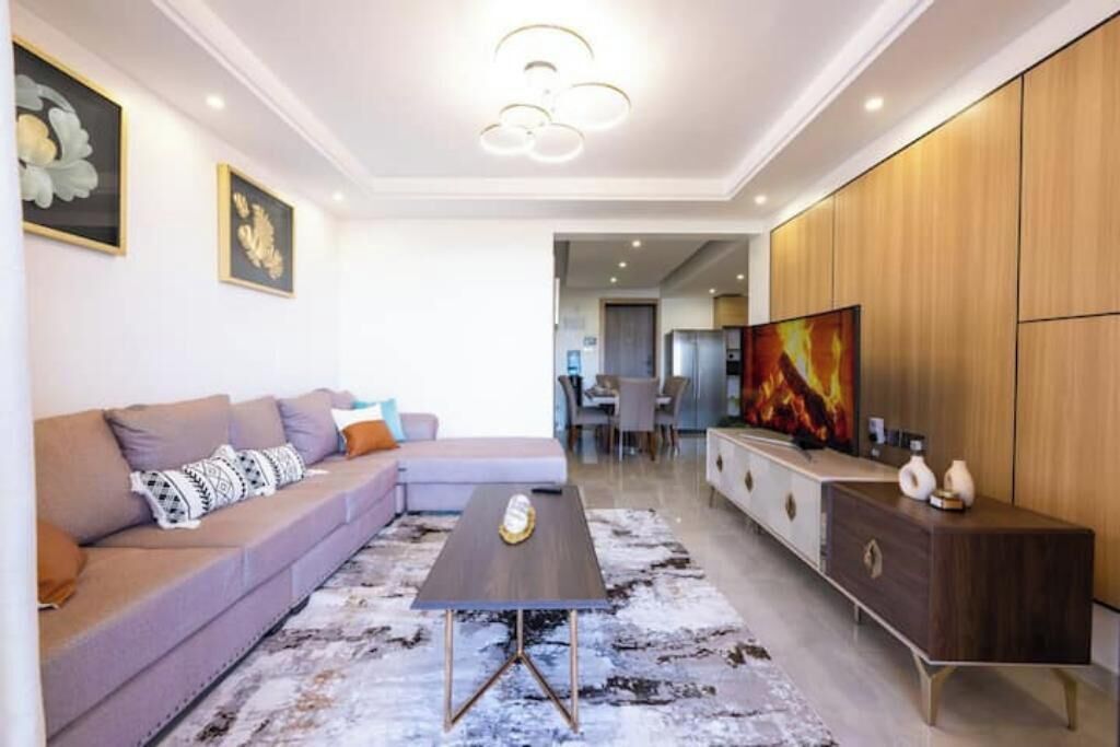 Un salon moderne avec un canapé, une table basse et un téléviseur. On aperçoit, au fond, un meuble en bois et un coin repas.